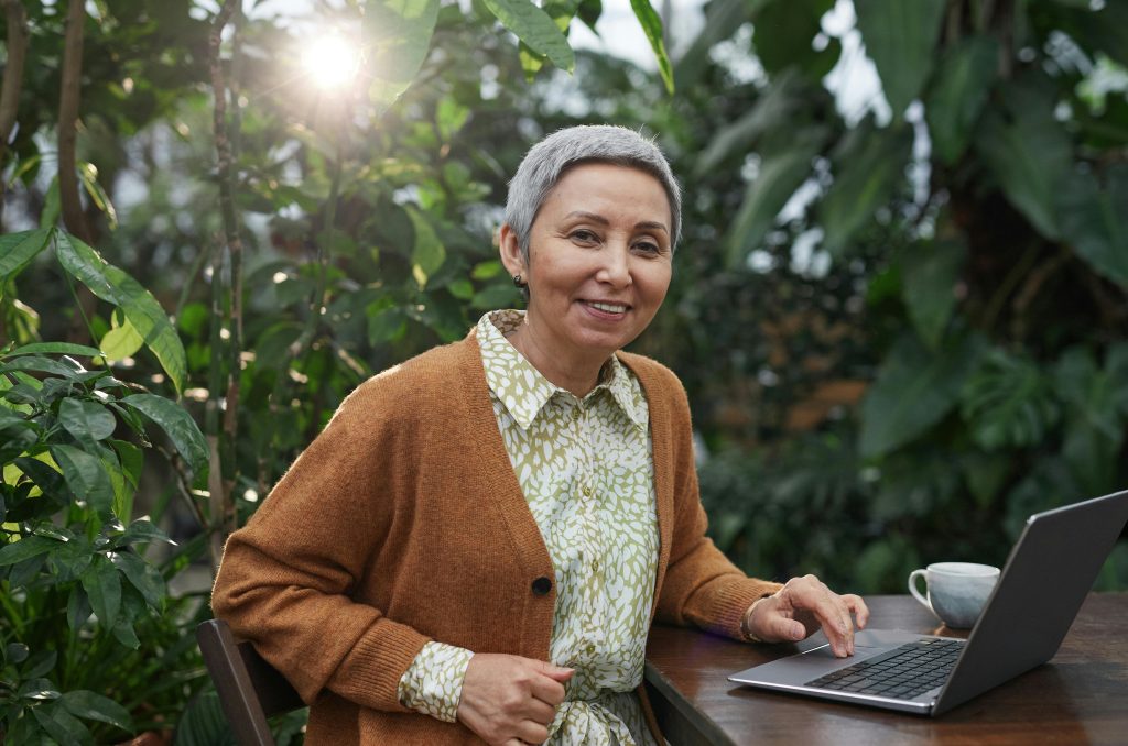 Senior woman smiling and using laptop in a lush botanical garden setting.
