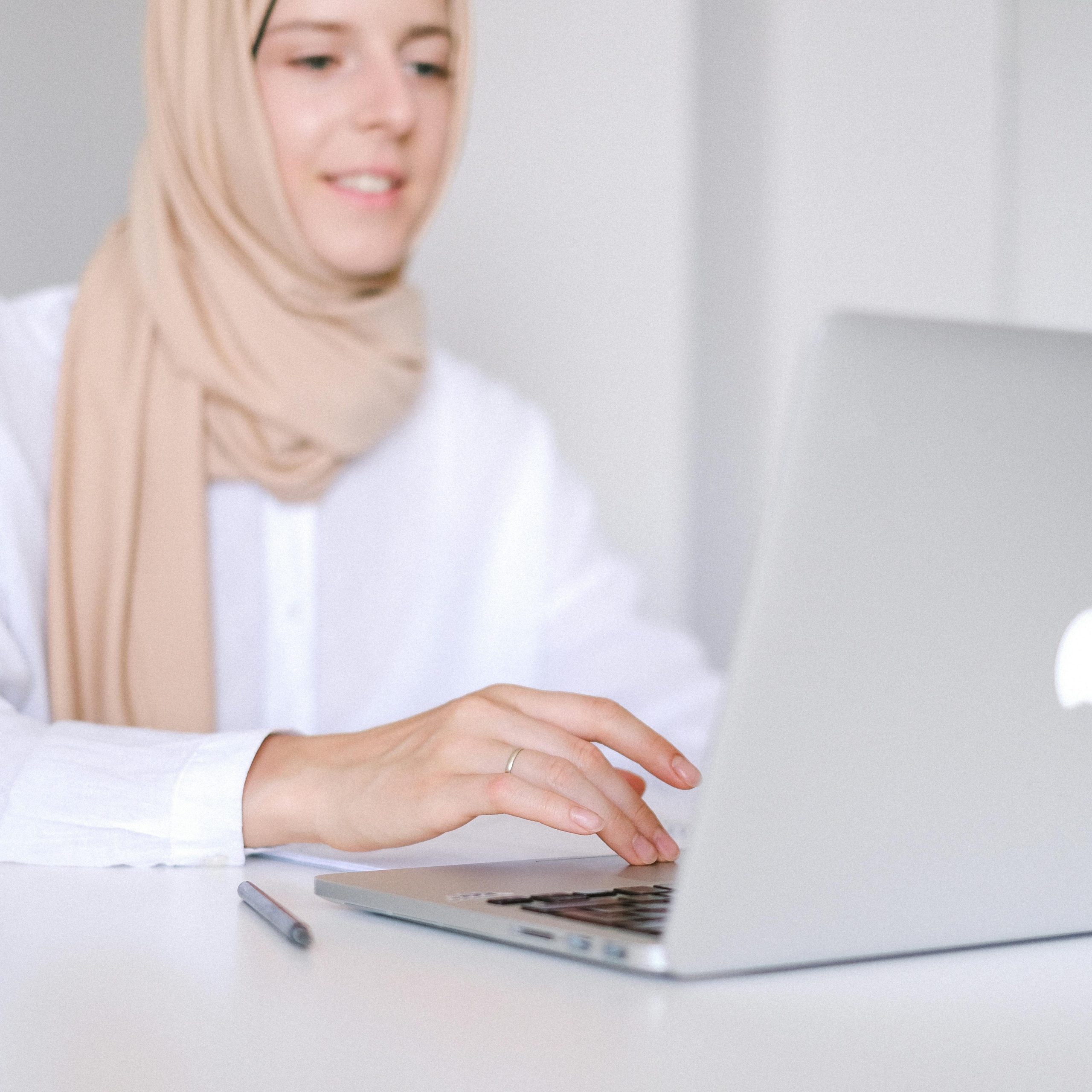 Muslim woman in headscarf smiling while working on laptop at home office.